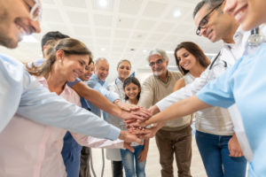 Group of healthcare workers and patients of different ages and ethnicities in a huddle all with hands in smiling at the hospital.