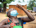 A woman in a rural area has her head tilted back as she holds a medicine tablet over her mouth. In her other hand she holds a tumbler of water.
