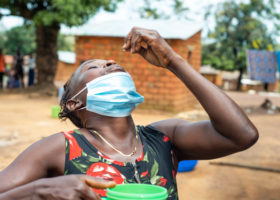 A woman in a rural area has her head tilted back as she holds a medicine tablet over her mouth. In her other hand she holds a tumbler of water.