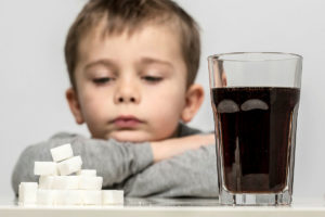 A young child rests their chin on their arms looking at a pile of sugar cubes and a glass of a carbonated cola drink on a table